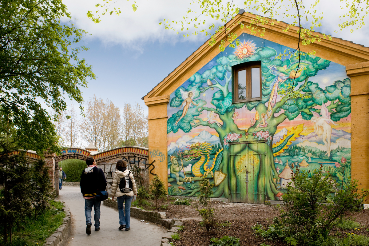 pareja paseando por el barrio de Christiania. En la casa se ve una casa con un mural pintado que representa un arbol y alrededor seres fantásticos como hadas, duendes y un dragón